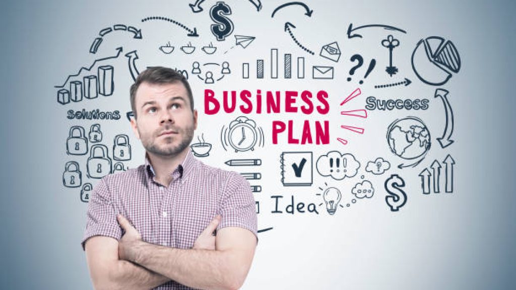 Portrait of a pensive young man wearing a checkered shirt and standing with crossed arms near a gray wall with a black and red business plan